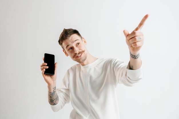 Young handsome man showing smartphone screen over gray background with a surprise face. Human emotions, facial expression concept