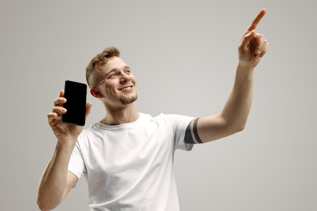 Young handsome man showing smartphone screen over gray background with a surprise face. Human emotions, facial expression concept