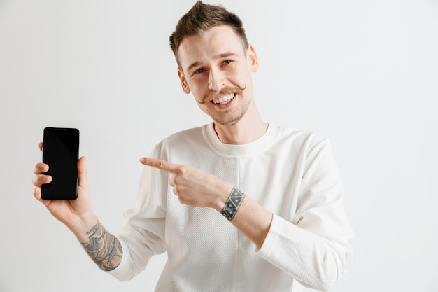 Young handsome man showing smartphone screen over gray space with a surprise face