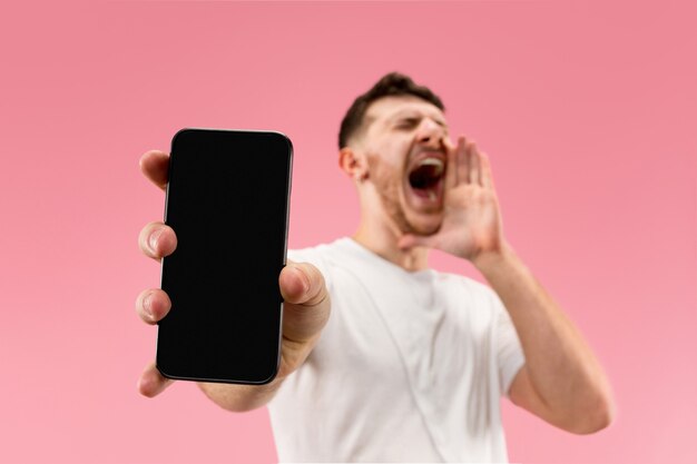 Young handsome man showing smartphone screen isolated on pink background in shock with a surprise