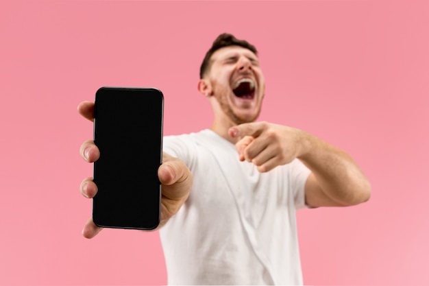 Young handsome man showing smartphone screen over pink background with a surprise face. Human emotions, facial expression concept. Trendy colors