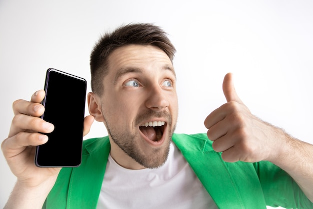 Young handsome man showing smartphone screen and signing OK sign isolated on gray background. Human emotions, facial expression, advertising concept.