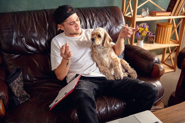 Free photo young handsome man sitting on brown sofa and working with his cute dog