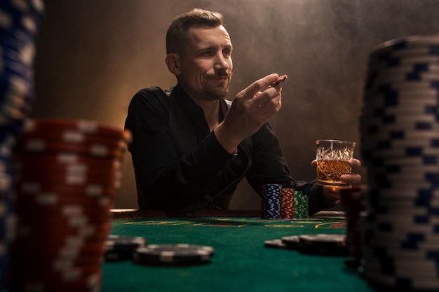 Free photo young handsome man sitting behind poker table with cards and chips. in the foreground stacks of chips on the poker table in a dark room full of cigarette smoke.