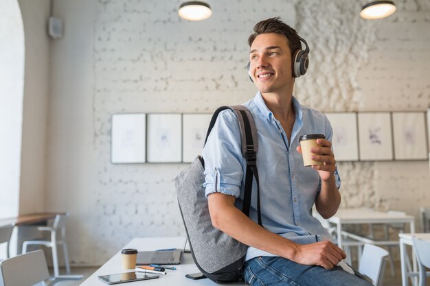 コーヒーを飲みながらコワーキングオフィスでバックパックとヘッドフォンのテーブルの上に座っている若いハンサムな男