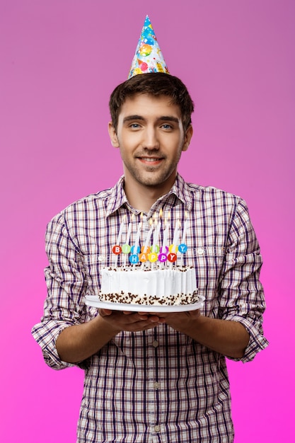 Young handsome man smiling, holding birthday cake over purple wall.