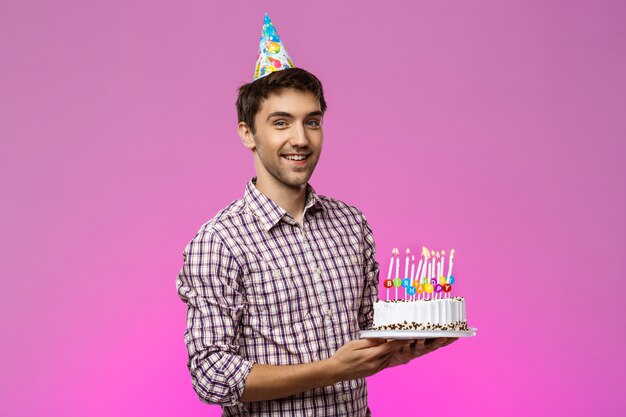 Молодой красавец улыбается, держа торт ко дню рождения над фиолетовым стены.