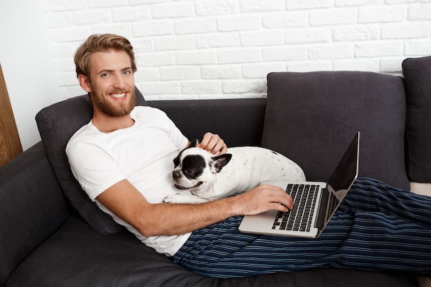 Young handsome man smiling holding laptop stroking pug dog.