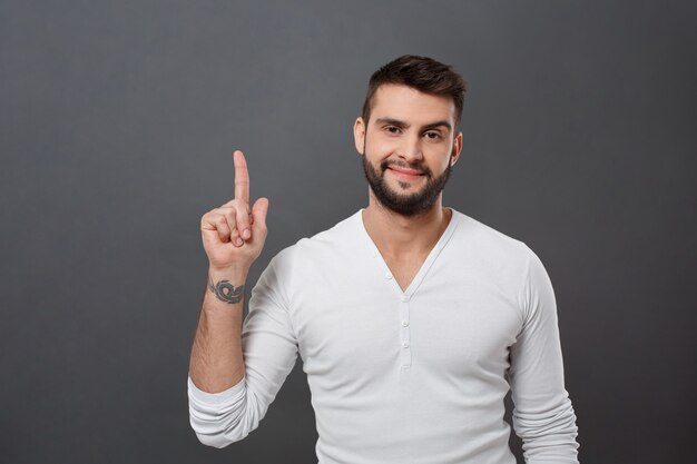 Young handsome man smiling pointing finger up over grey wall