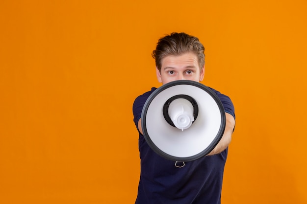 Young handsome man speaking to megaphone looking at camera positive and happy standing over orange background