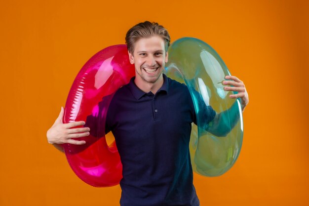 Young handsome man standing with inflatable rings looking at camera happy and exited smiling cheerfully over orange background