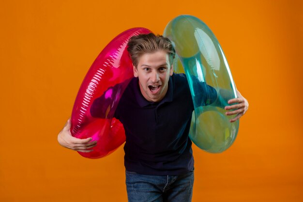 Young handsome man standing with inflatable rings surprised and happy with wide open mouth over orange background