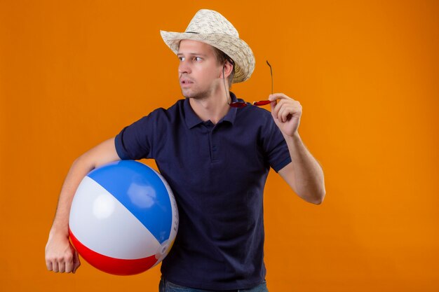 Young handsome man in summer hat holding inflatable ball and sunglasses looking aside with serious face uncertain look standing over orange background