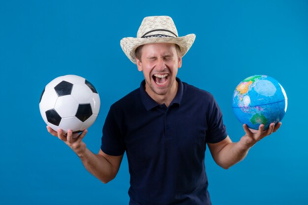 Young handsome man in summer hat holding soccer ball and globe crazy happy screaming in fascination standing over blue background