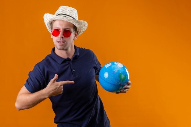 Free photo young handsome man in summer hat wearing red sunglasses holding globe pointing finger to it with confident expression on face standing over orange background