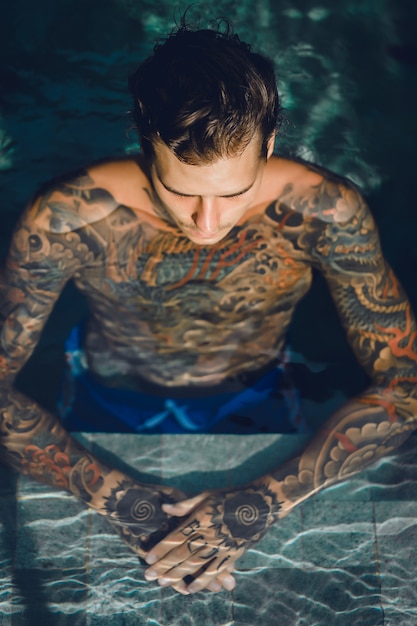 Free photo young handsome man in tattoos resting in the outdoor pool.