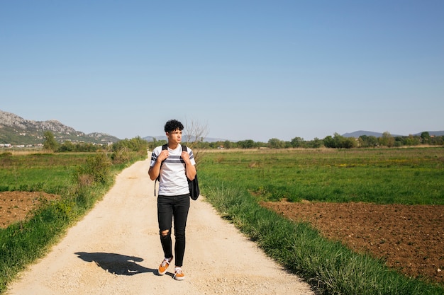 バックパックを運ぶ未舗装の道路の上を歩く若いハンサムな男