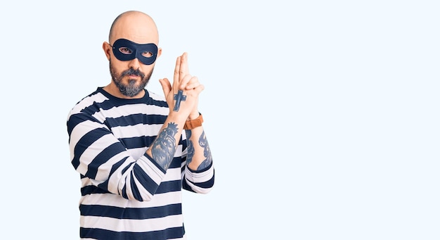 Young handsome man wearing burglar mask holding symbolic gun with hand gesture, playing killing shooting weapons, angry face