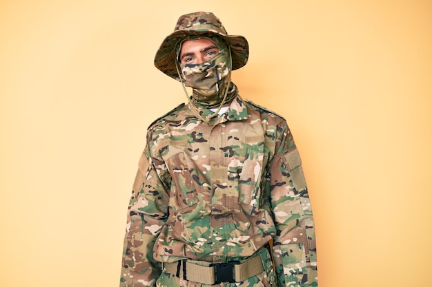 Free photo young handsome man wearing camouflage army uniform and balaclava looking positive and happy standing and smiling with a confident smile showing teeth