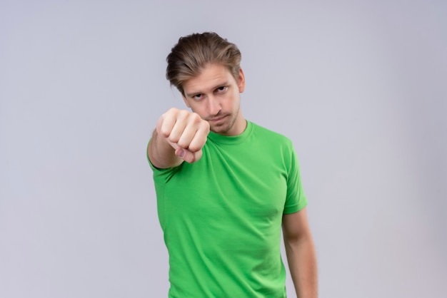 白い壁に深刻な顔立ってカメラに伸ばして緑のtシャツを食いしばって拳を身に着けている若いハンサムな男