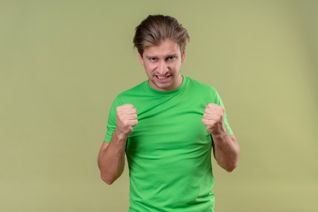 Free photo young handsome man wearing green t-shirt clenching fists with agressive expression standing over green wall