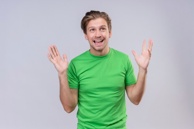 Young handsome man wearing green t-shirt crazy happy with raised arms standing over white wall 2