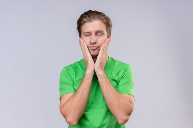 Giovane uomo bello che indossa la maglietta verde che tiene il viso con le mani che sembrano stanche in piedi sopra il muro bianco