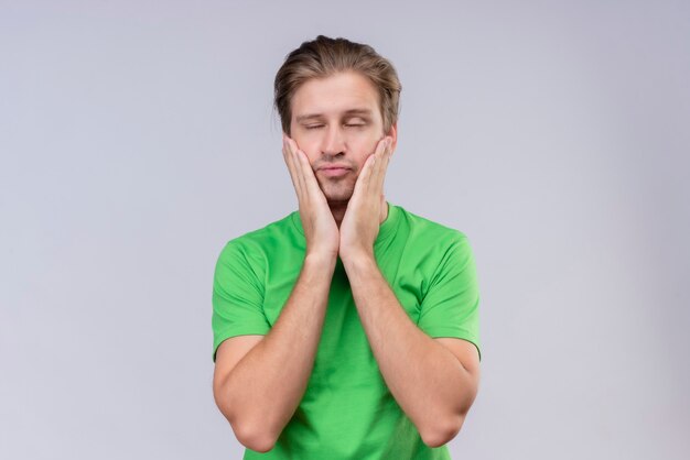 白い壁に疲れて立っているを探している手で彼の顔を止める緑のtシャツを着ている若いハンサムな男