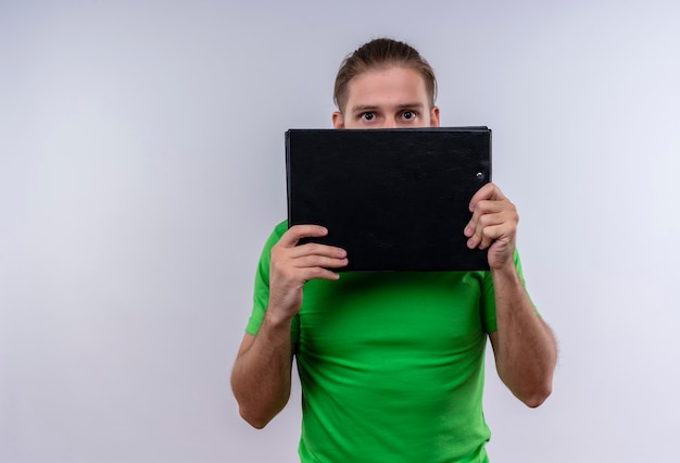 Young handsome man wearing green t-shirt holding document case hiding behind it standing over white background