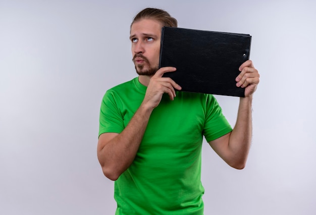 Giovane uomo bello che indossa la t-shirt verde tenendo la custodia del documento cercando di pensare in piedi su sfondo bianco