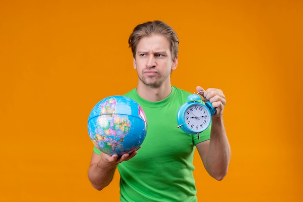 Free photo young handsome man wearing green t-shirt holding globe and alarm clock looking aside with frowning face displeased standing over orange wall