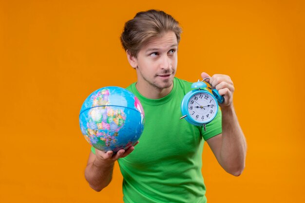 지구본과 알람 시계를 들고 녹색 티셔츠를 입고 젊은 잘 생긴 남자는 오렌지 벽에 옆으로 서있는 교활하게보고 웃고