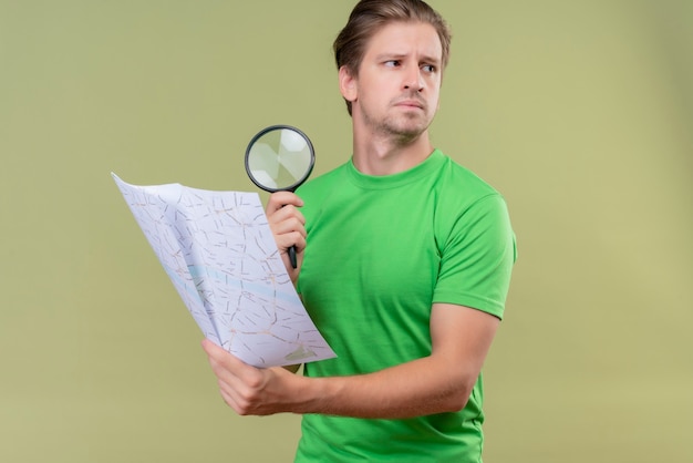 Giovane uomo bello che indossa t-shirt verde che tiene mappa e lente di ingrandimento con espressione seria sul viso che osserva da parte in piedi sopra la parete verde