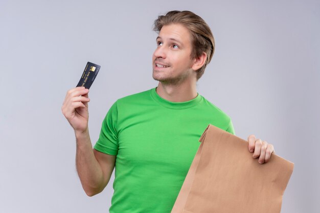 Young handsome man wearing green t-shirt holding paper package and credit car looking up