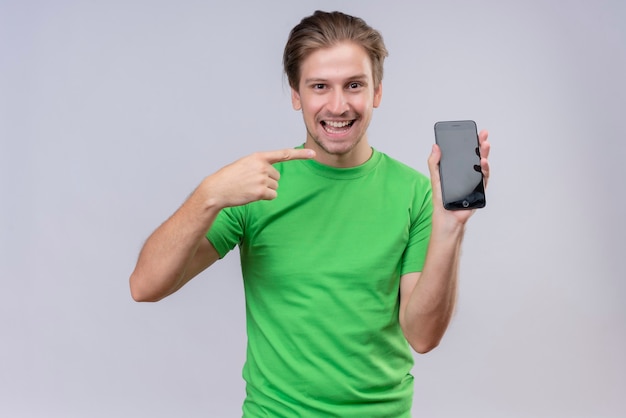 Молодой красавец в зеленой футболке держит и показывает смартфон, указывая пальцем на него, весело улыбаясь, стоя над белой стеной