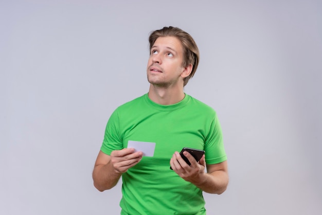 スマートフォンとクレジットカードを保持している緑のtシャツを着ている若いハンサムな男が白い壁に立って選択をしようと考えて顔の物思いに沈んだ表情で見上げる