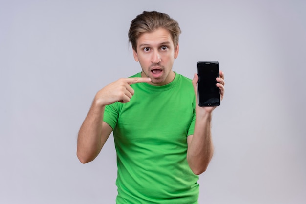 Young handsome man wearing green t-shirt holding smartphone pointing with finger to it looking surprised and confused standing over white wall