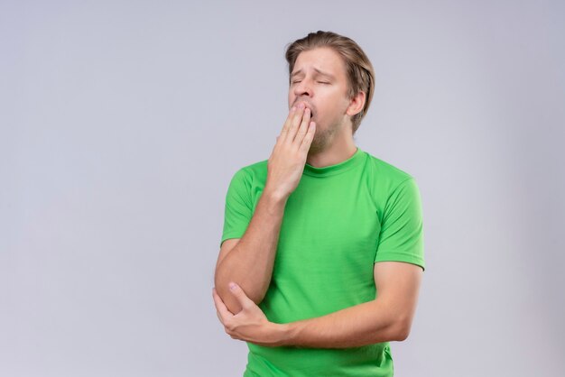 Young handsome man wearing green t-shirt looking tired yawning standing over white wall