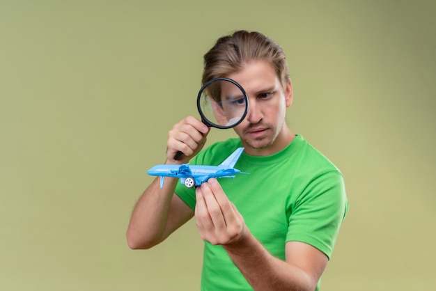 緑の壁の上に立っている顔に真剣な表情で虫眼鏡を通しておもちゃの飛行機を見て緑のtシャツを着ている若いハンサムな男