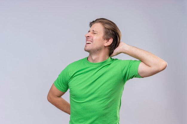 Young handsome man wearing green t-shirt looking unwell feeling pain in back standing over white wall