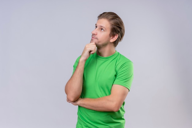 Foto gratuita giovane uomo bello che indossa la maglietta verde che osserva in su con la mano sul mento con l'espressione pensierosa sul fronte che sta sopra il muro bianco