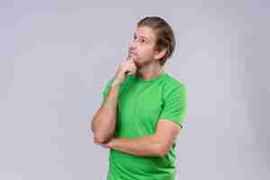 Free photo young handsome man wearing green t-shirt looking up with hand on chin with pensive expression on face standing over white wall