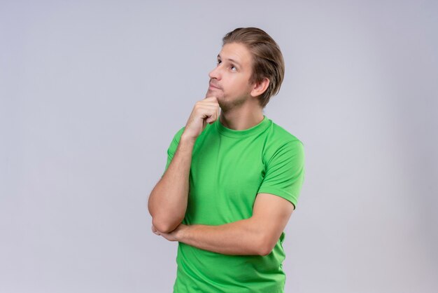 흰 벽 위에 서있는 얼굴에 잠겨있는 표정으로 턱에 손을 올려 녹색 티셔츠를 입고 젊은 잘 생긴 남자