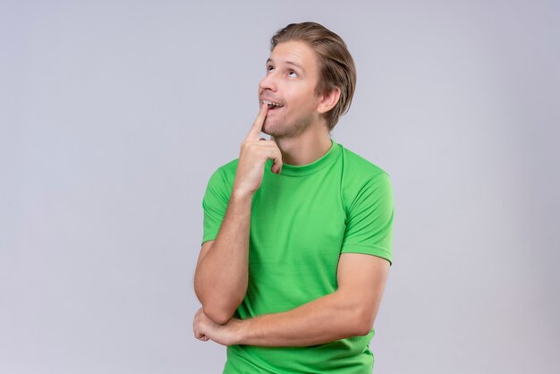 Young handsome man wearing green t-shirt looking up with pensive expression thinking positive standing over white wall