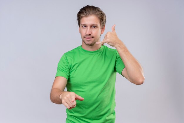 Young handsome man wearing green t-shirt making call me gesture smiling friendly standing over white wall