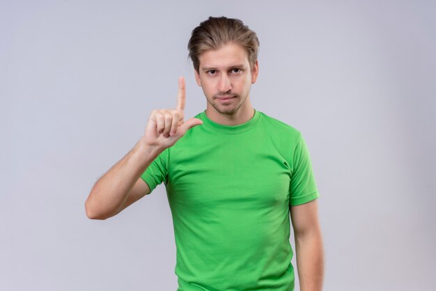 Молодой красивый мужчина в зеленой футболке, указывая пальцем вверх с уверенным выражением лица, стоит над белой стеной