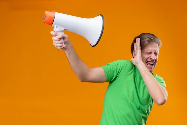 Young handsome man wearing green t-shirt protecting with hand against megaphone with closed eyes annoyed standing over orange wall
