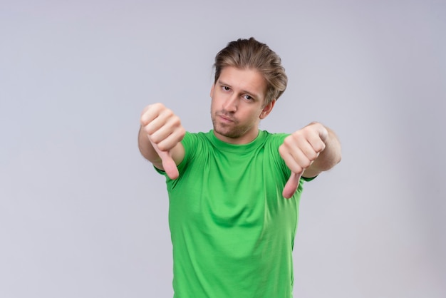 Young handsome man wearing green t-shirt unhappy showing thumbs down standing over white wall