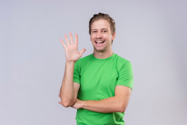 흰 벽 위에 서있는 손으로 흔들며 녹색 티셔츠를 입고 젊은 잘 생긴 남자