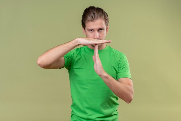 Foto gratuita giovane uomo bello che indossa la maglietta verde con la faccia accigliata che fa il gesto di time out con le mani in piedi sopra la parete verde